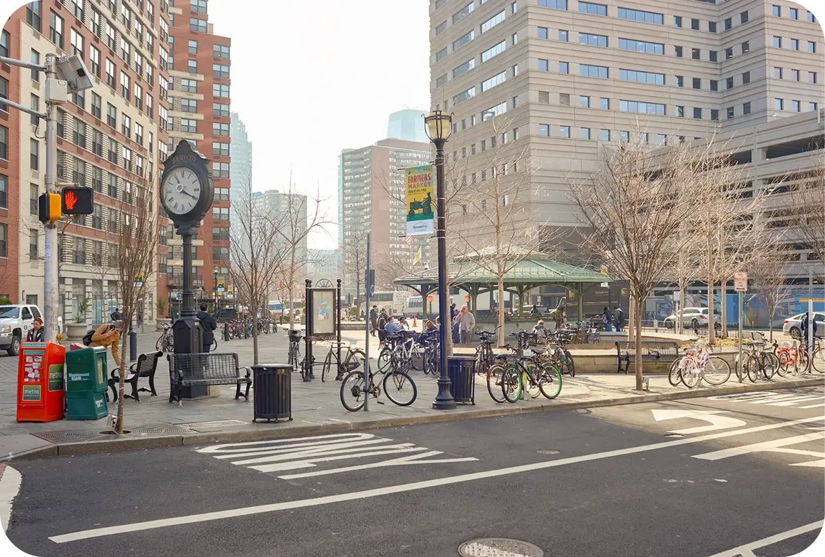 Urban plaza in Jersey City with people, bicycles, a vintage clock, and tall buildings