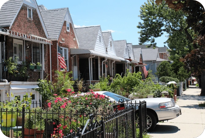 Row of charming brick and siding houses with front gardens and American flags in a residential Queens neighborhood, with a car parked along the sidewalk