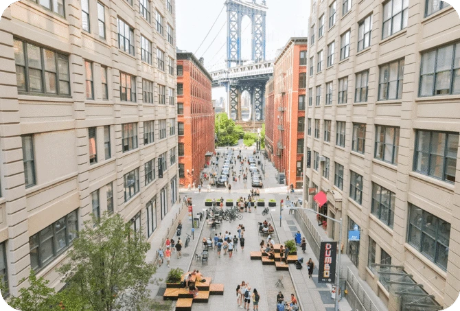 View of the Manhattan Bridge framed by buildings in DUMBO, Brooklyn, with people walking and sitting at outdoor tables